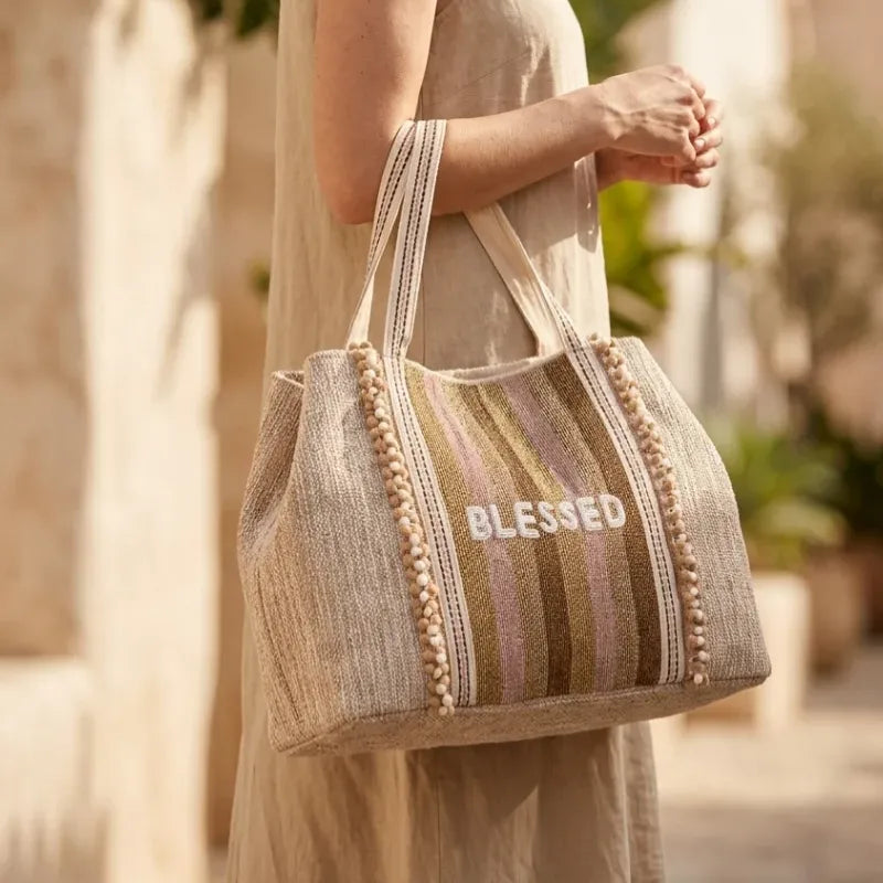 Beige woven tote bag with striped pattern pom-pom trim and blessed embroidery ideal for women’s boho chic clothing shop daisy