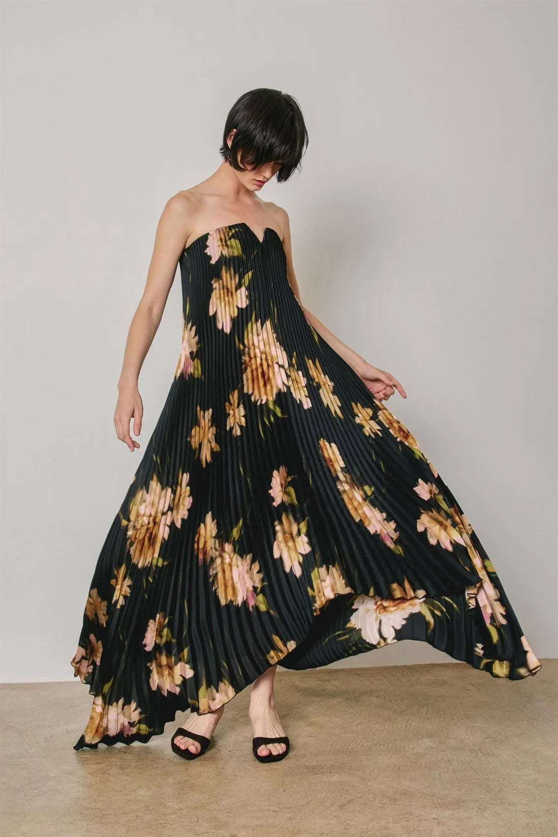 A black floral strapless pleated maxi column dress featuring vibrant peach and gold floral patterns in soft pink tones