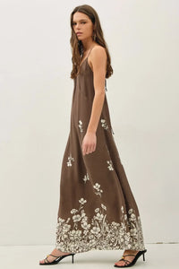 A flowing brown maxi dress with delicate white floral printed strappy tie details along the hemline and straps