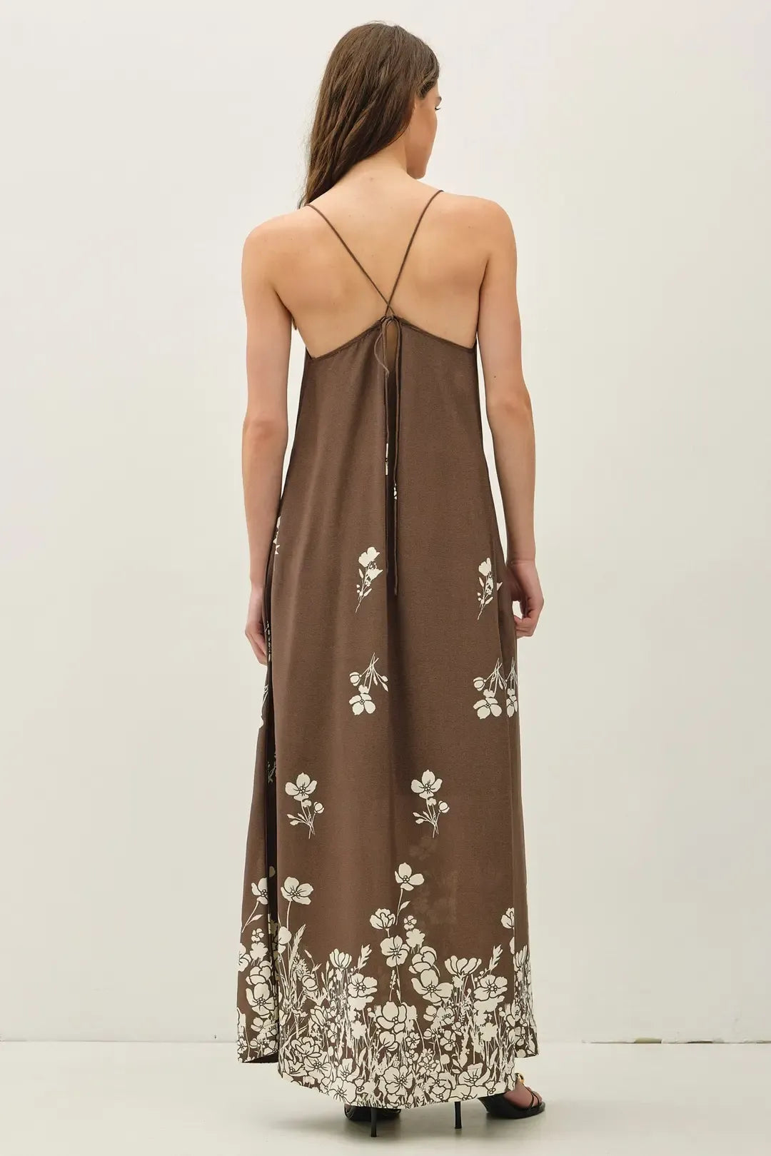A flowing brown maxi dress with delicate white floral embroidery and thin spaghetti straps crossing at the neckline