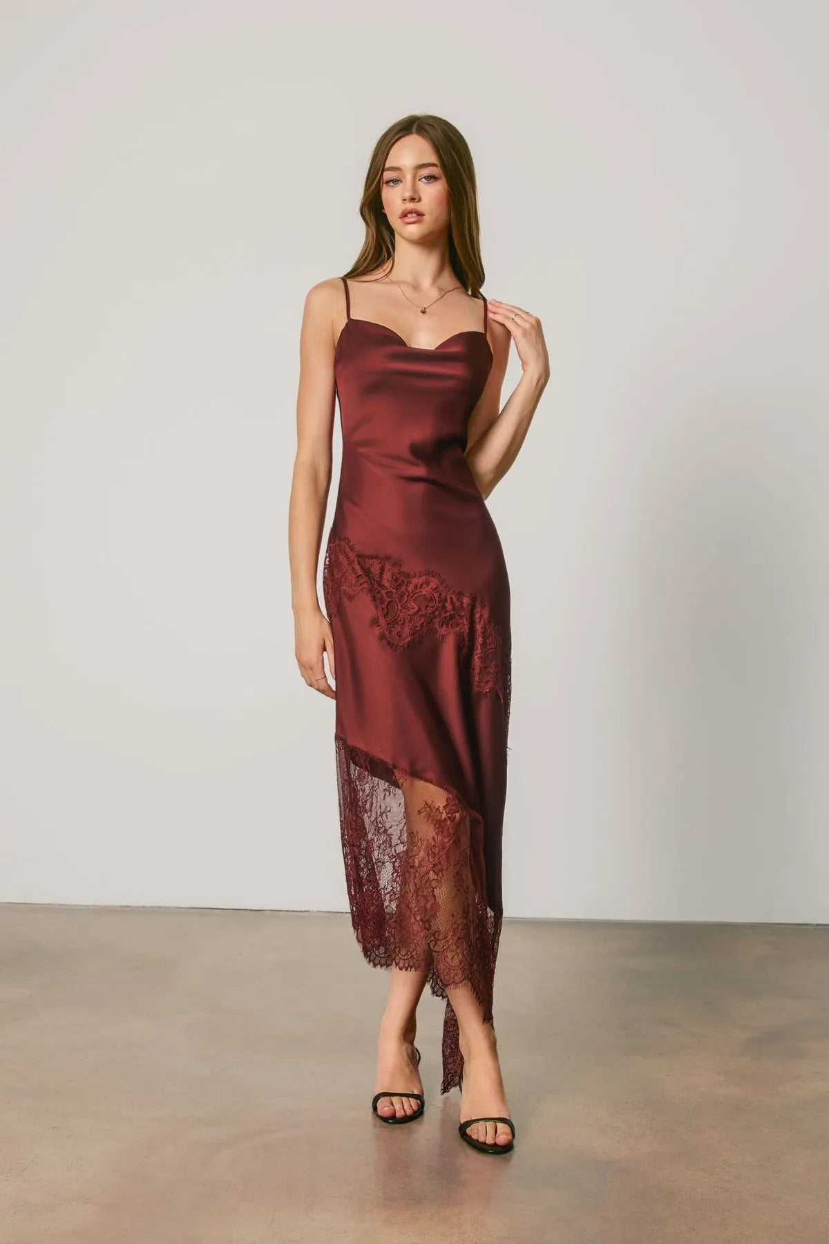A flowing burgundy satin midi slip dress with asymmetrical lace trim along the hem and side slit