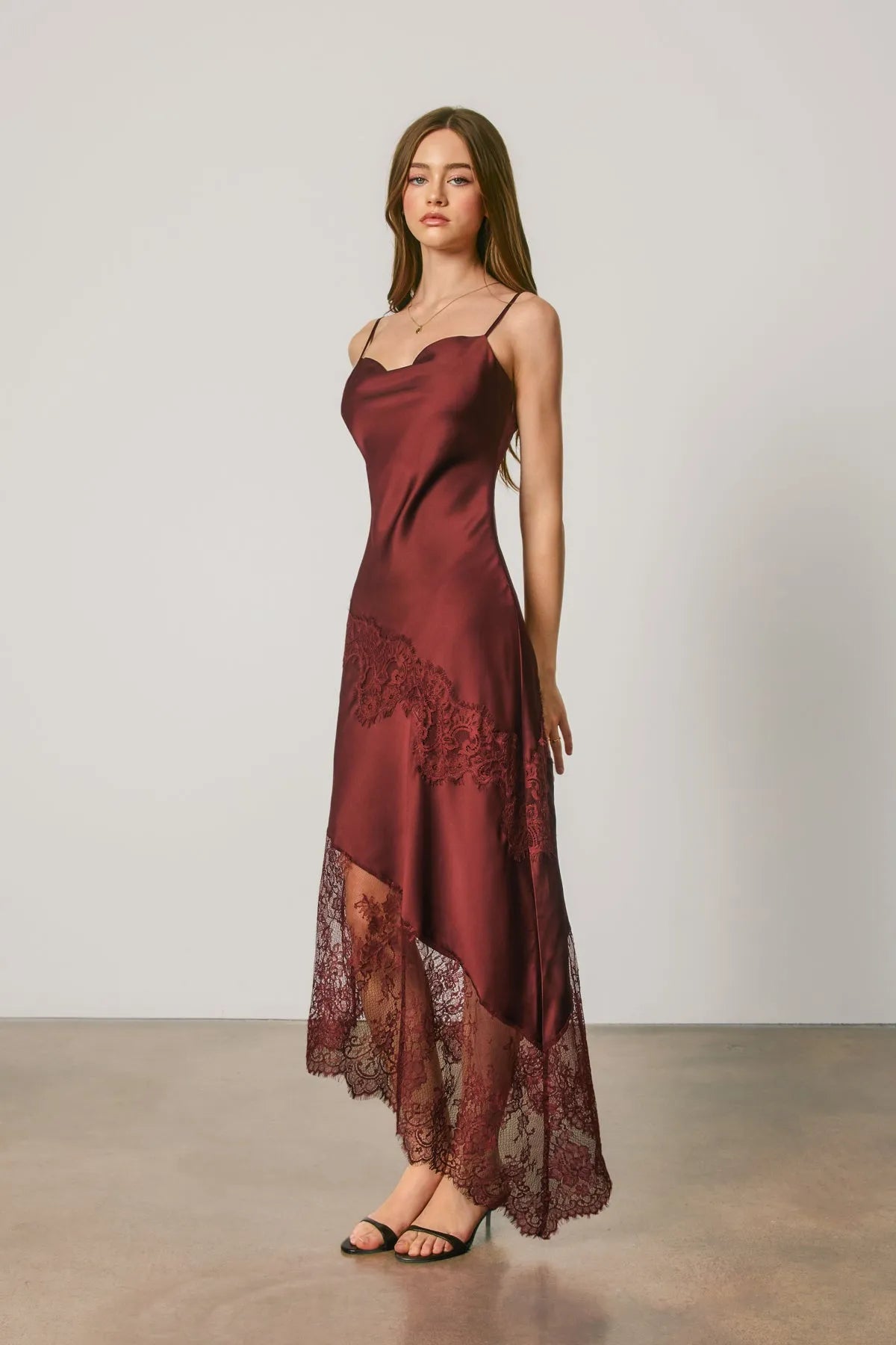 A flowing burgundy satin slip dress with asymmetrical lace trim along the hem and side panels