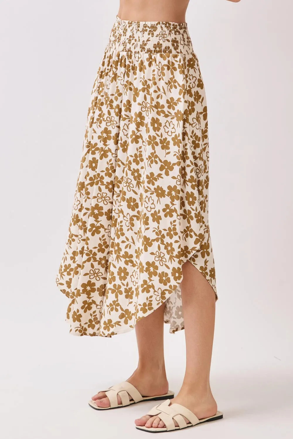 A cream-colored smocked waistband midi skirt with a gold floral pattern and an asymmetrical hemline