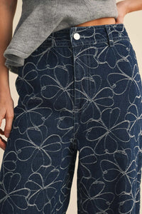 Dark blue denim pants with a white floral design distressed hem featuring a repeating four-leaf clover pattern