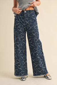 Dark blue denim wide-leg pants with white floral design and distressed hem featuring frayed edges