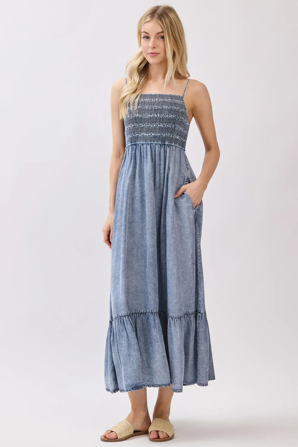 A denim blue tencel smocked tiered midi dress featuring a sleeveless maxi design with a smocked bodice and tiered skirt