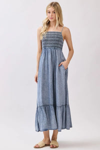 A denim blue tencel smocked tiered midi dress featuring a sleeveless maxi design with a smocked bodice and tiered skirt
