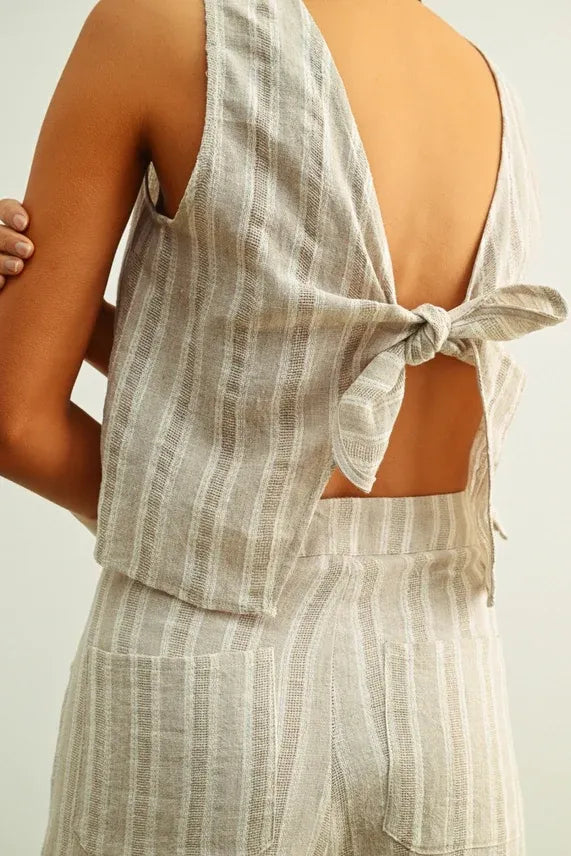 A light gray and white vertically striped jacquard linen twisted tie back jumpsuit shown from the back side