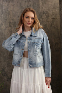 Light-wash oversized trucker bow patch denim jacket with a decorative bow appliqué on the left chest pocket