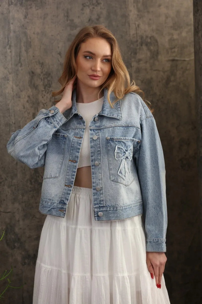 Light-wash oversized trucker bow patch denim jacket with a decorative bow appliqué on the left chest pocket