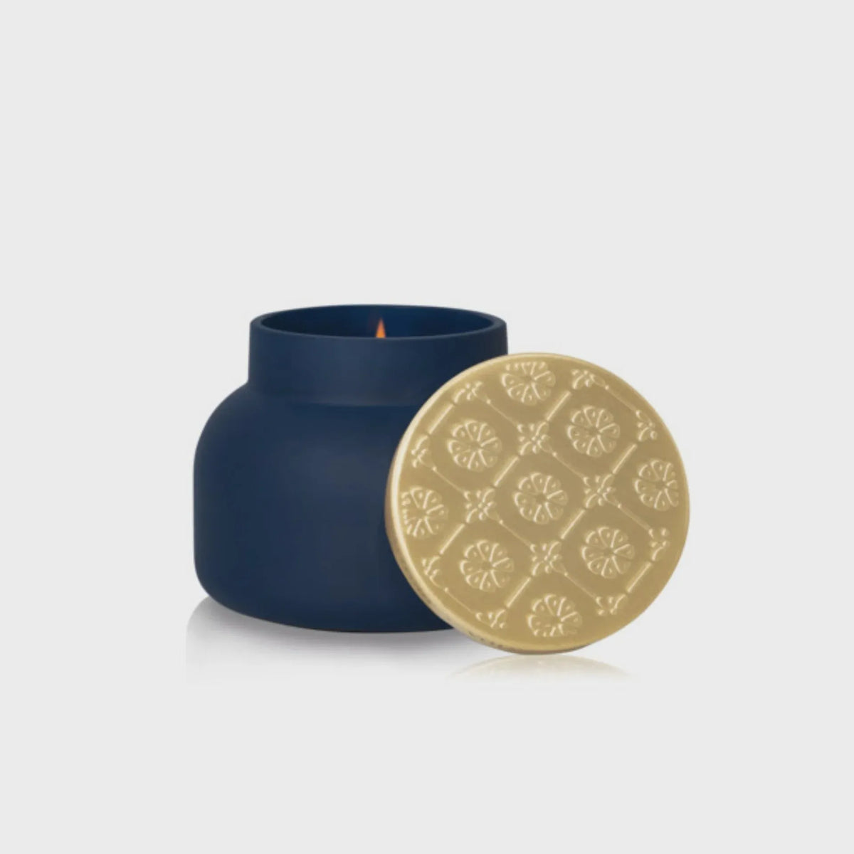 A navy blue ceramic candle jar with a gold embossed lid and softly glowing flame from capri blue artisan terrazza velluto
