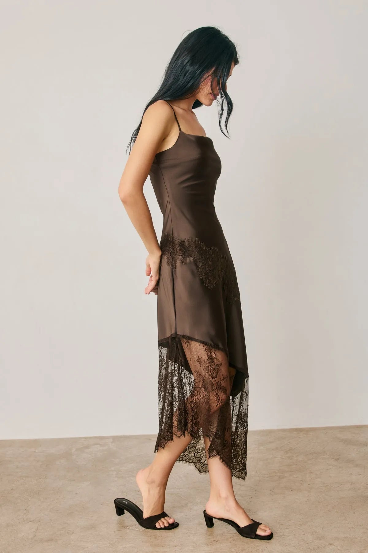 A sleek dark brown satin midi slip dress with asymmetrical black lace trim along the hemline