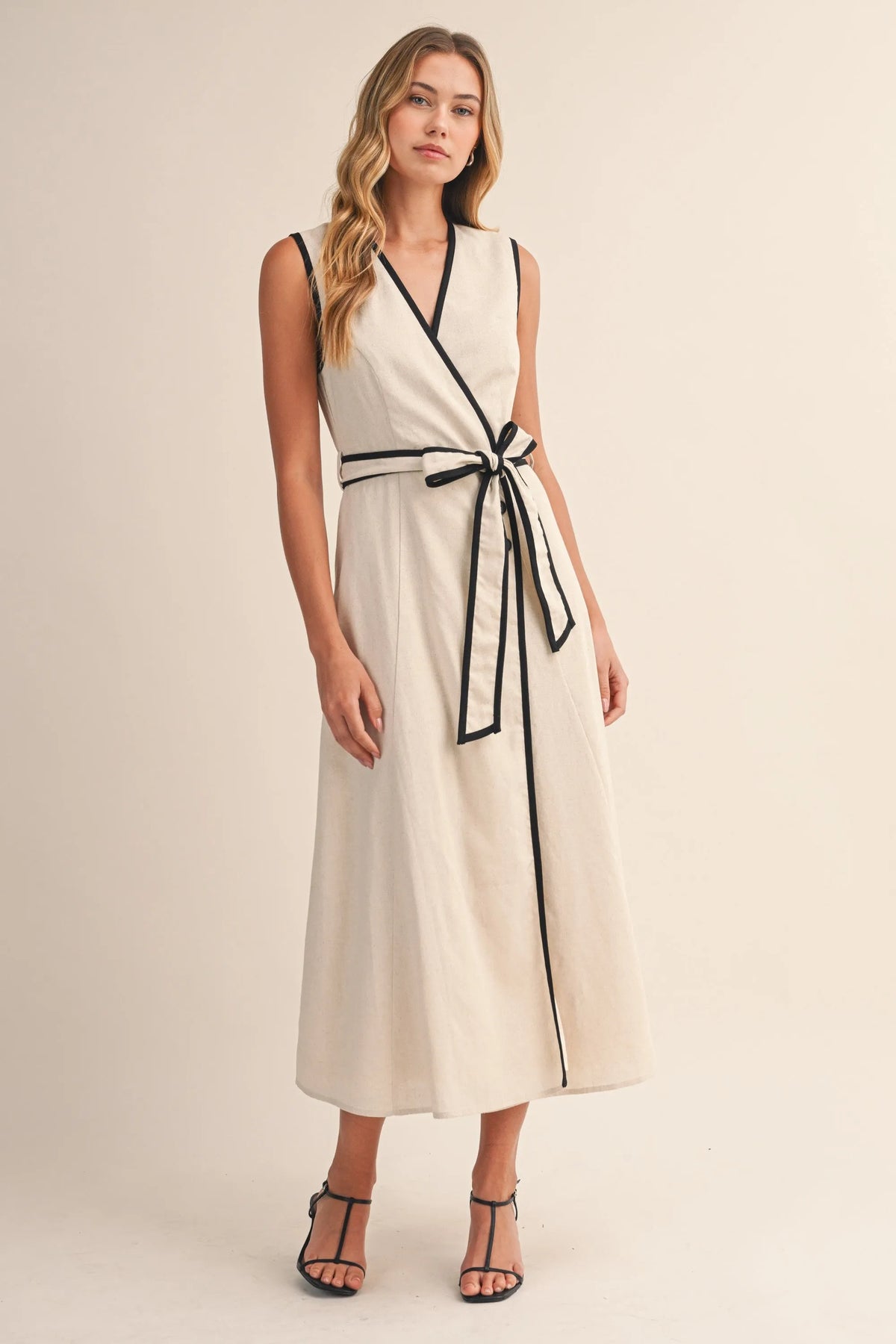 A sleeveless cream midi dress with black contrast piping and a matching belt in a wrap midi dress style