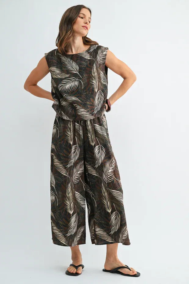 A flowing sleeveless jumpsuit with a dark brown base and white leaf print wide shoulder design