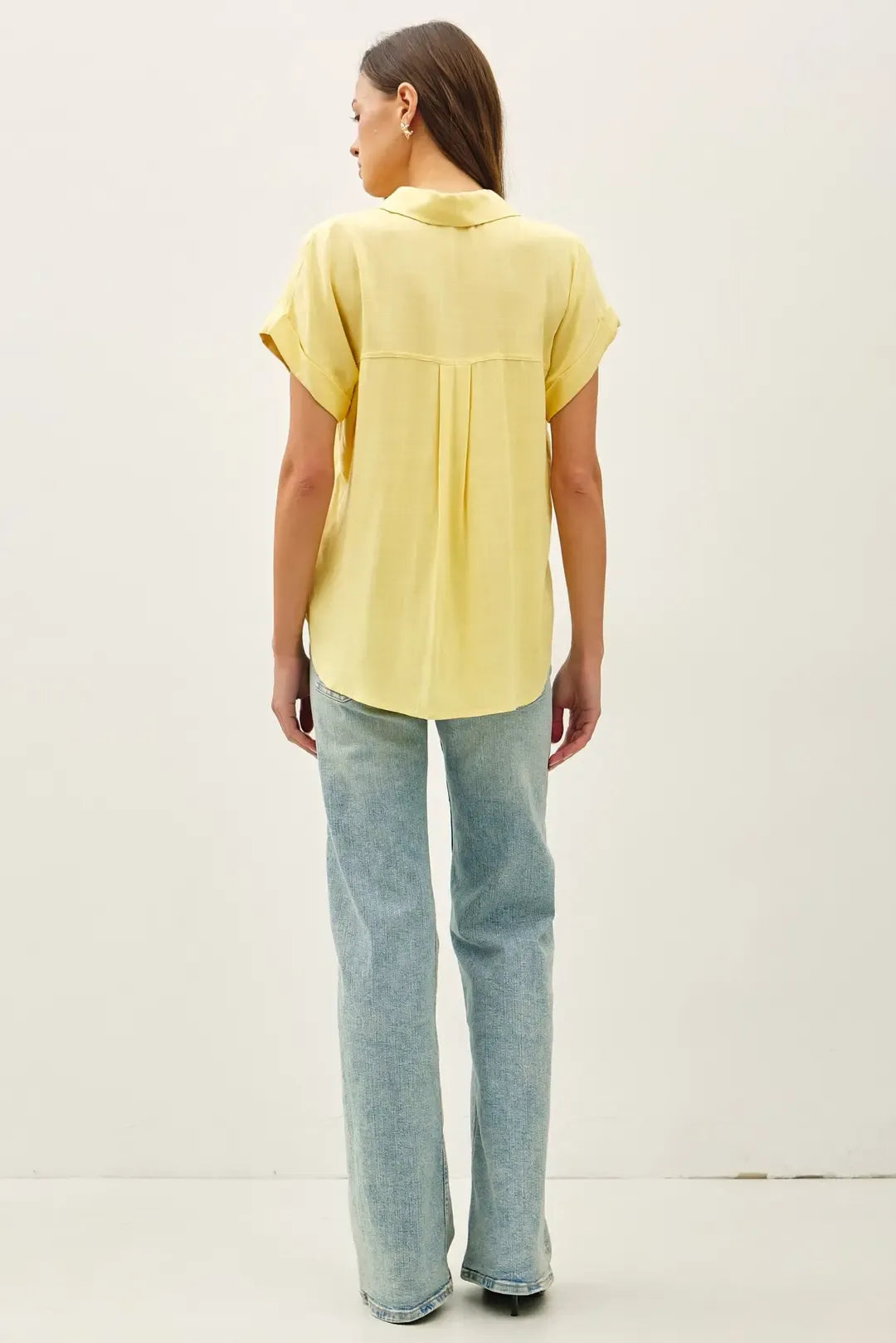 A soft yellow solid short sleeve button down high low shirt with relaxed fit and subtle back pleats shown from the back