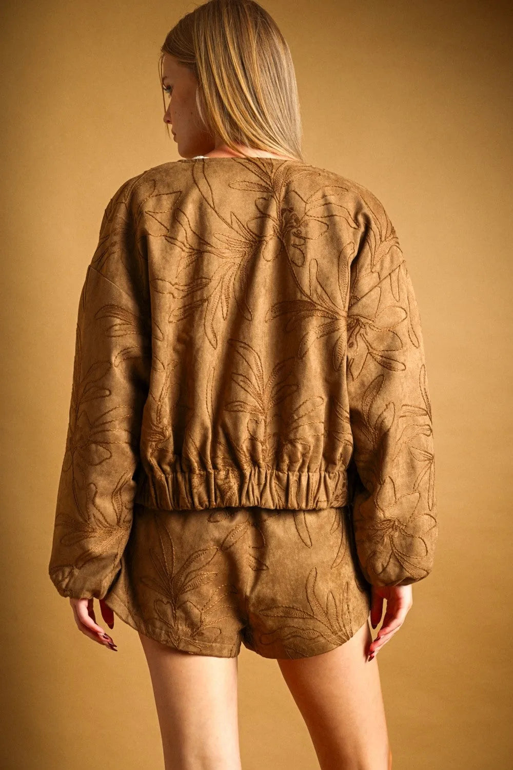 Tan suede bomber jacket with embroidered leaf pattern complementing suede floral textured shorts design