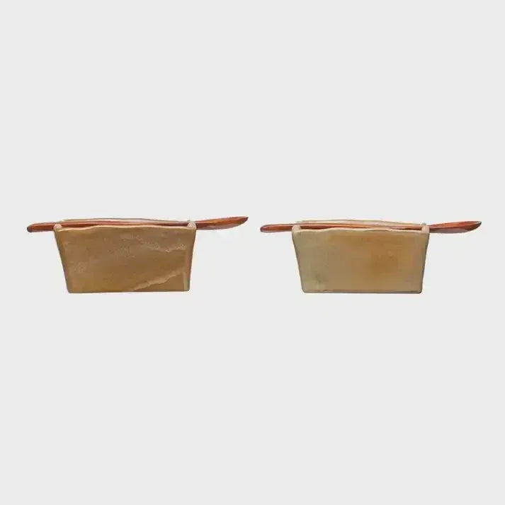 Two beige stoneware salt and pepper shakers with wooden lids beside a handcrafted wooden spoon on a stoneware dish