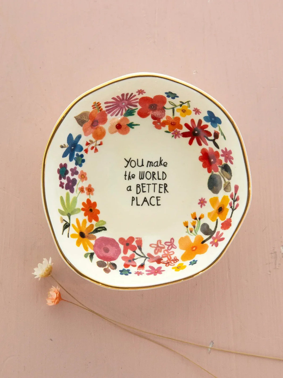 A white ceramic trinket bowl with a gold rim and colorful floral wreath featuring you make the world a better place inscription