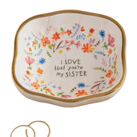 A white ceramic antiqued trinket bowl with gold rim and colorful floral patterns inscribed i love that youre my sister