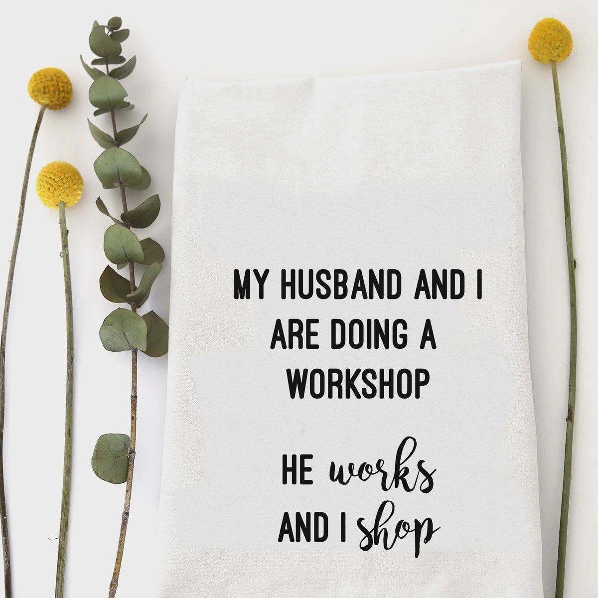 White cotton workshop tea towel with humorous black text: My husband and I are doing a workshop he works and I shop