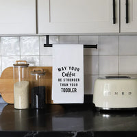 White kitchen tea towel with black text: May your coffee be stronger than your toddler