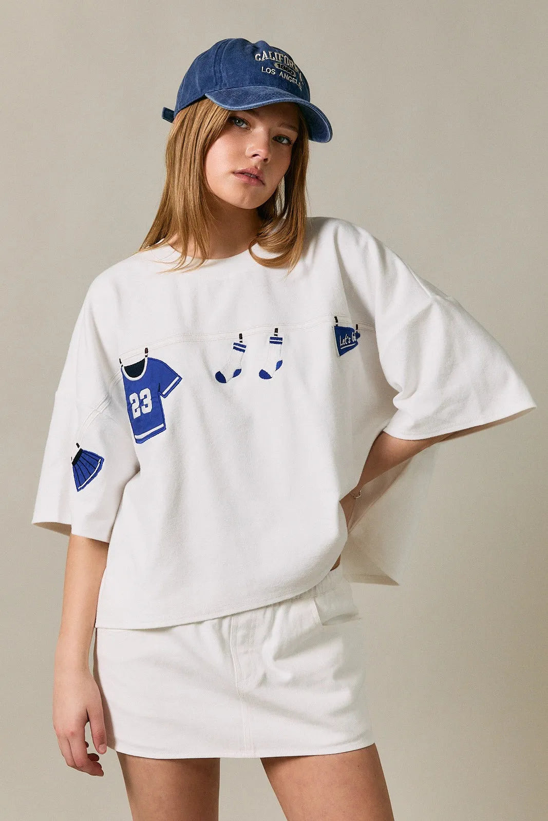 White oversized short sleeve americana top with blue sports-themed appliqué patches of jersey socks and pom-poms