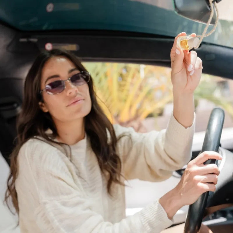 A woman holds a clear glass bottle car diffuser with a golden interior hanging air freshener for luxury scent