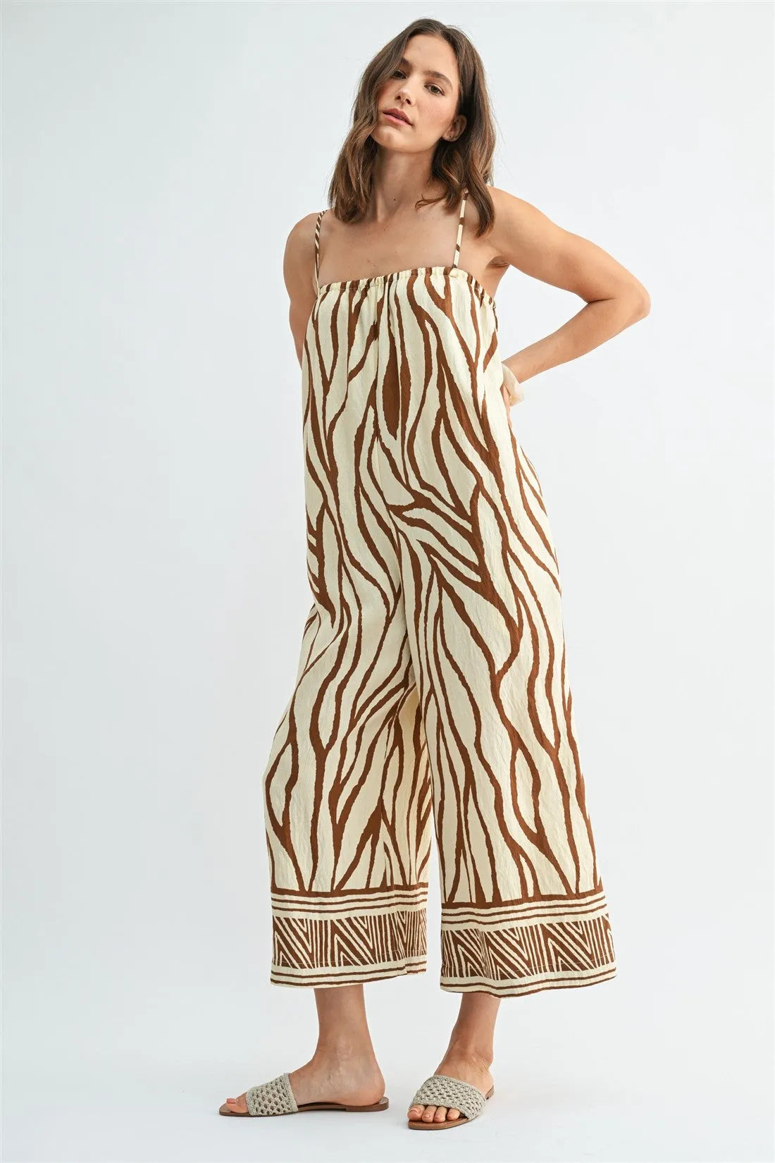 A woman wears a beige and brown border print jumpsuit with wide legs and geometric patterned cuffs