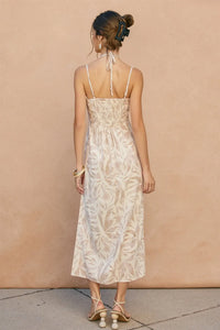 A woman wearing a beige tropical leaf sleeveless maxi dress with thin straps and a back tie detail