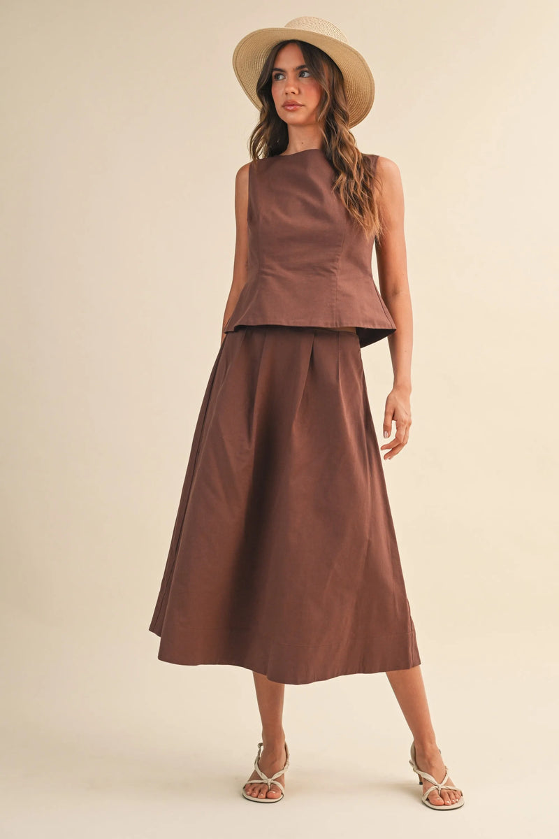 A woman wears a brown linen pleated waist flare midi skirt with a sleeveless top featuring structured peplum detailing