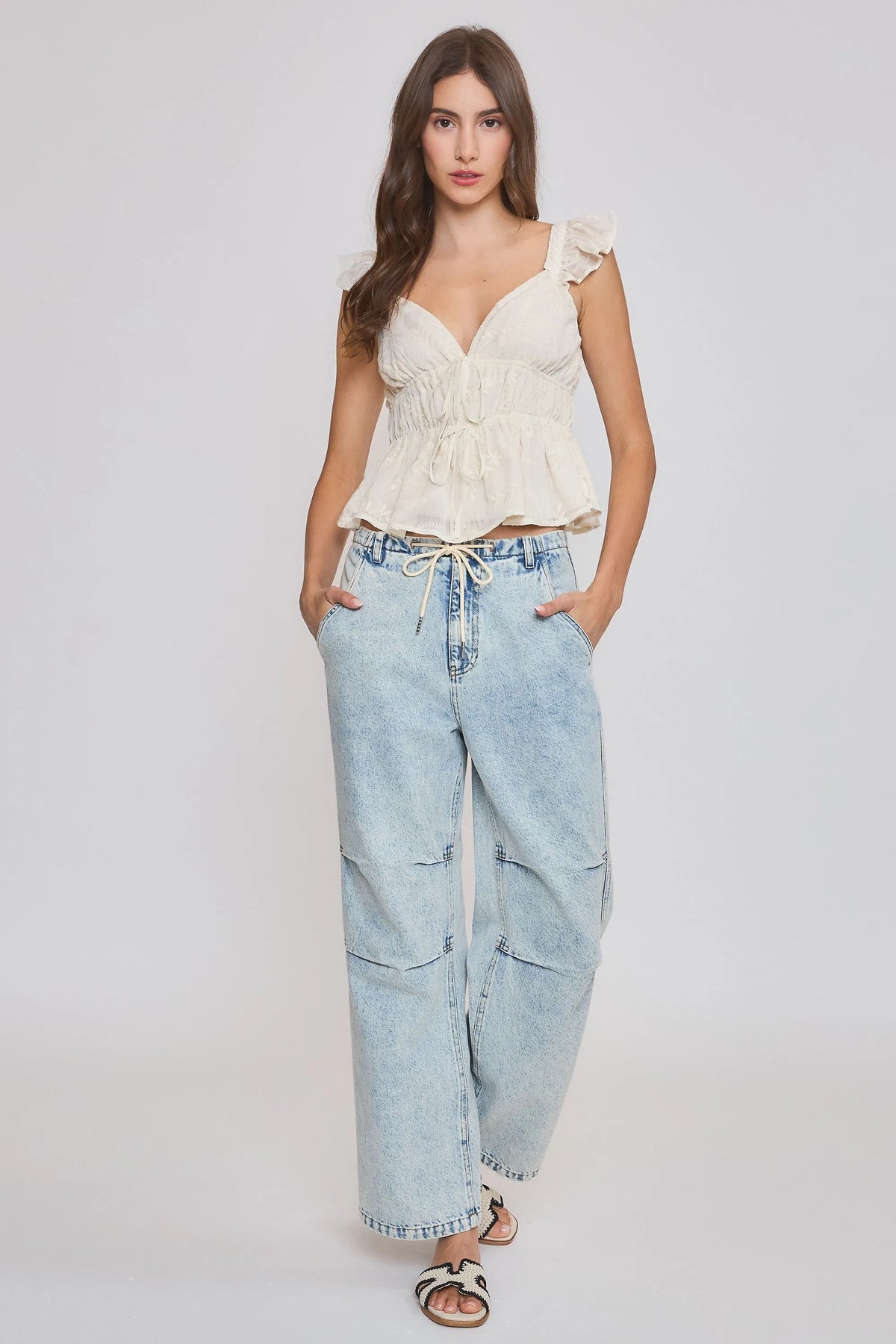 A woman wears a cream-colored embroidered ruffle sleeve blouse with thin straps and light-wash denim pants