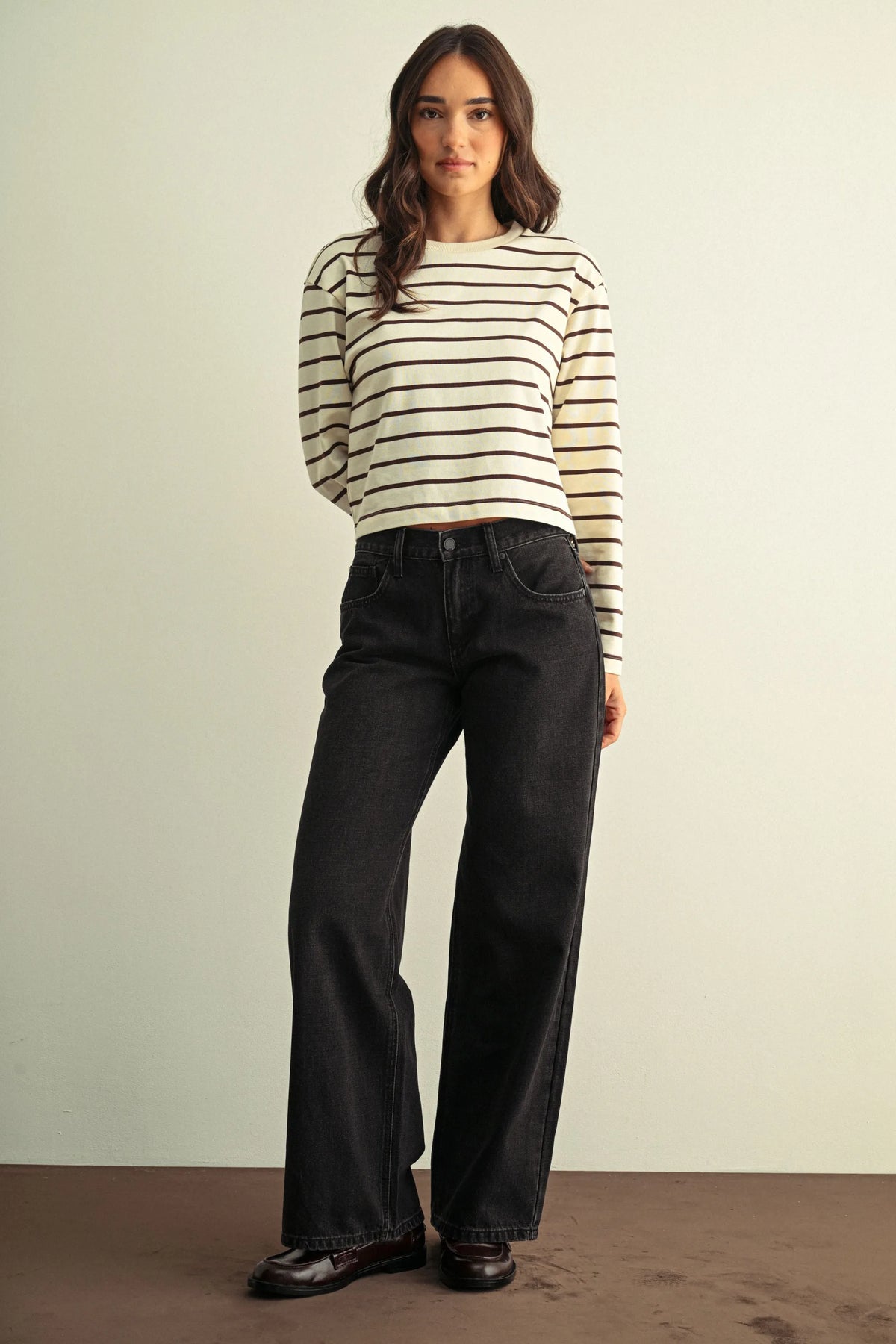 A woman wears low rise baggy wide dark denim jeans paired with a cream and black striped long-sleeved sweater