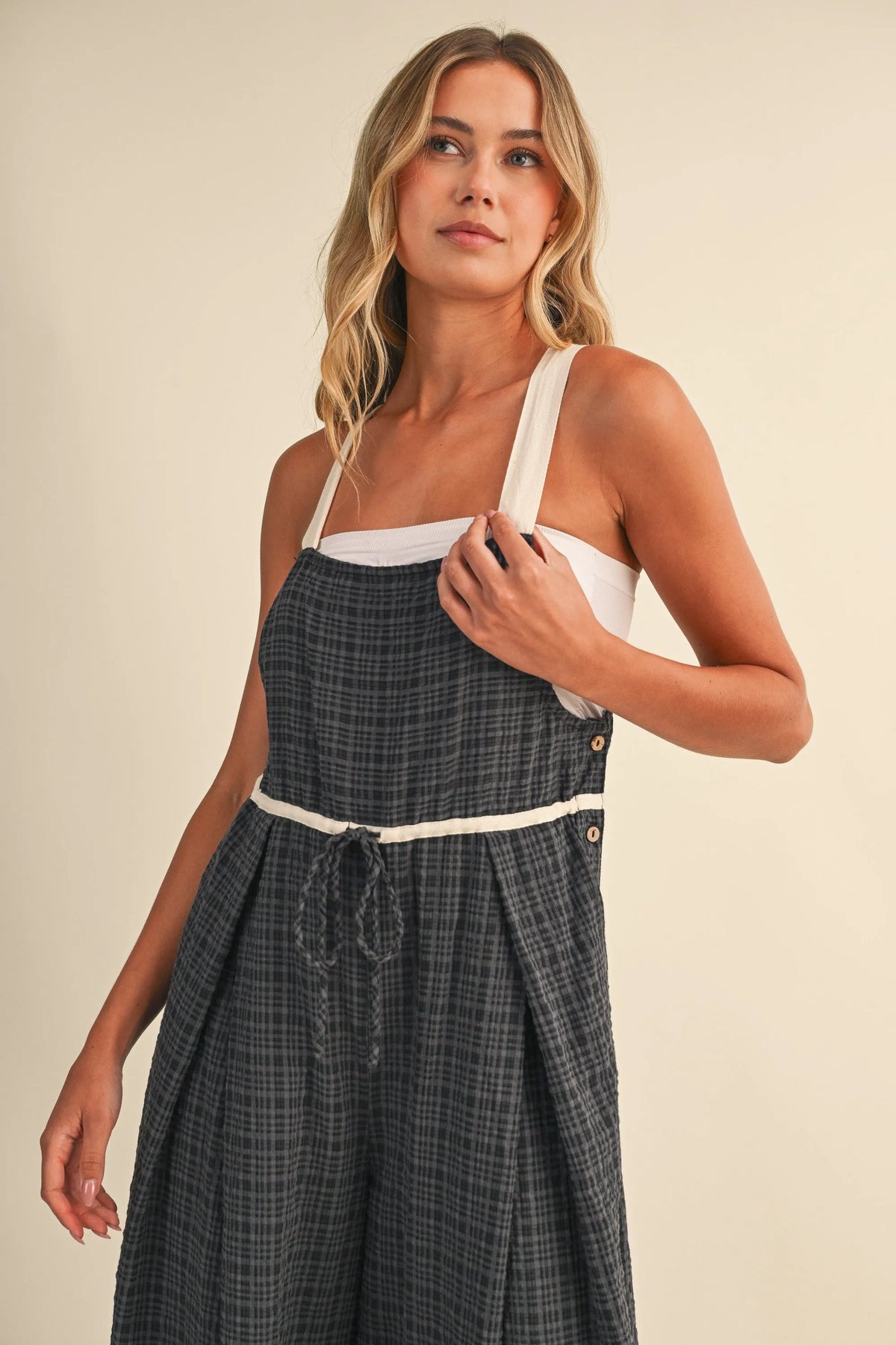 A woman wears a dark gray drawcord waistline plaid suspender dress with white straps trim and adjustable waistline plaid features