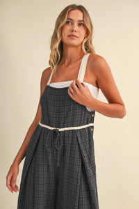 A woman wears a dark gray drawcord waistline plaid suspender dress with white straps trim and adjustable waistline plaid features