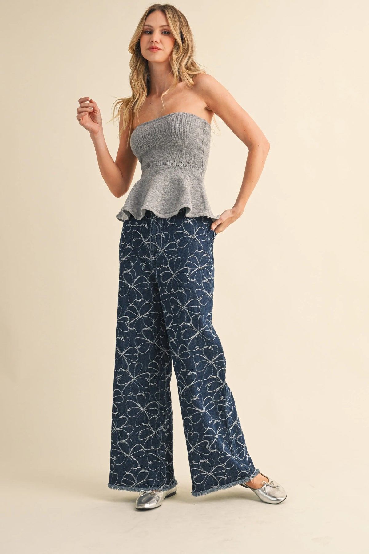 A woman wears navy blue denim pants with white floral design distressed hem paired with a gray peplum top