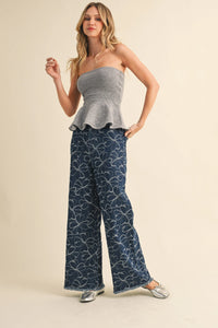 A woman wears navy blue denim pants with white floral design distressed hem paired with a gray peplum top