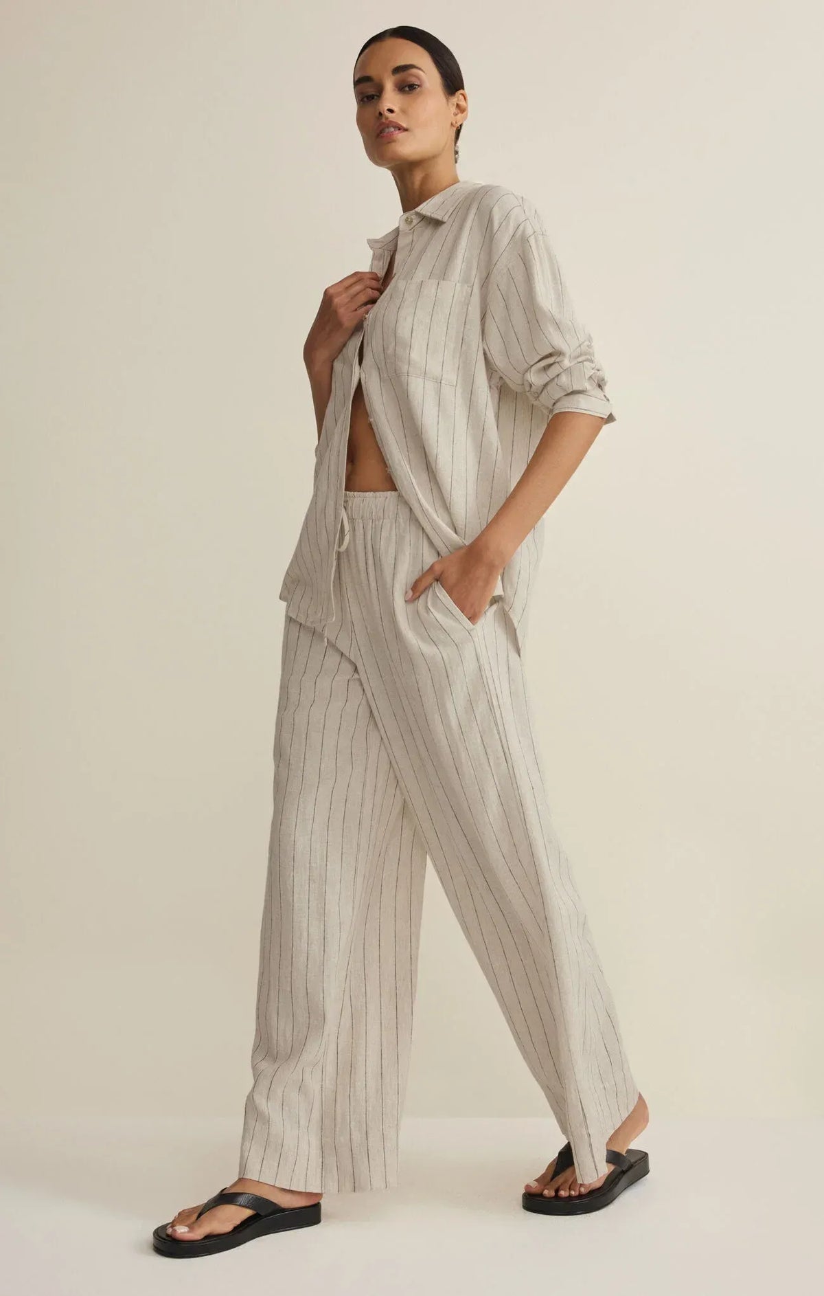 A woman wearing a henri striped wide light gray and white linen pajama set with striped wide leg pants and an oversized shirt