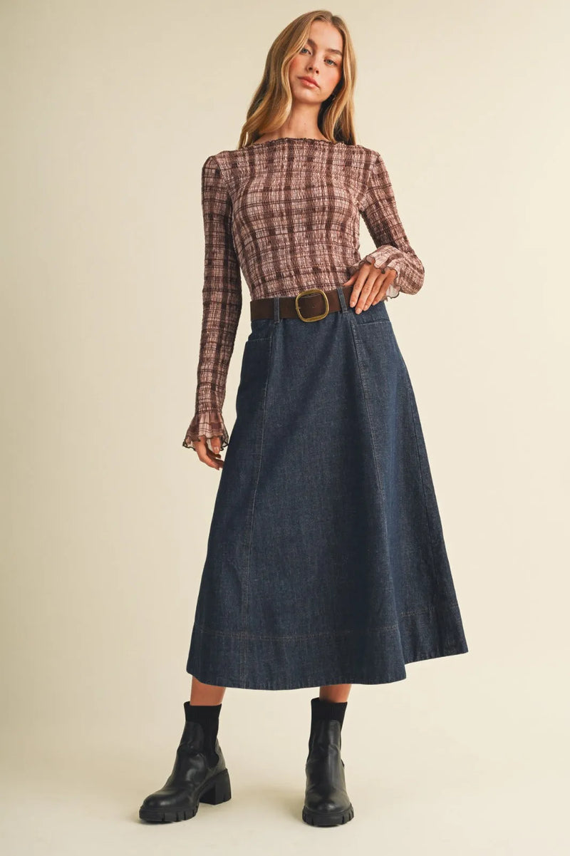 A woman wearing a belted indigo maxi skirt paired with a long-sleeved brown and beige plaid top with flared cuffs