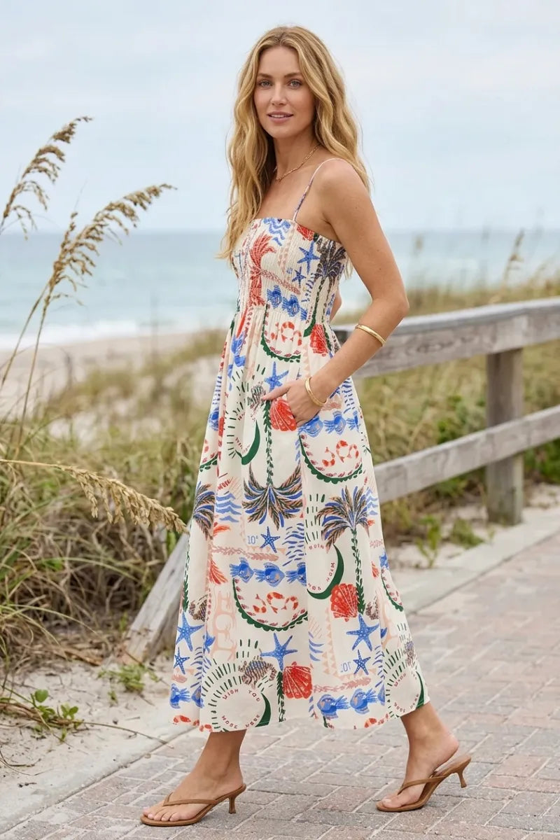 A woman wears a flowing off-white getaway maxi dress with vibrant blue starfish red coral and green palm fronds pattern