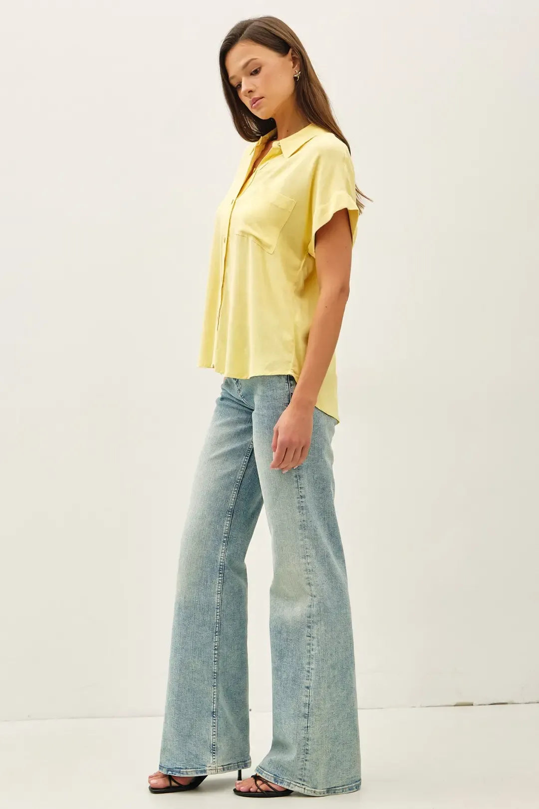 A woman wears a pale yellow solid short sleeve button down high low shirt with light-wash denim flare jeans