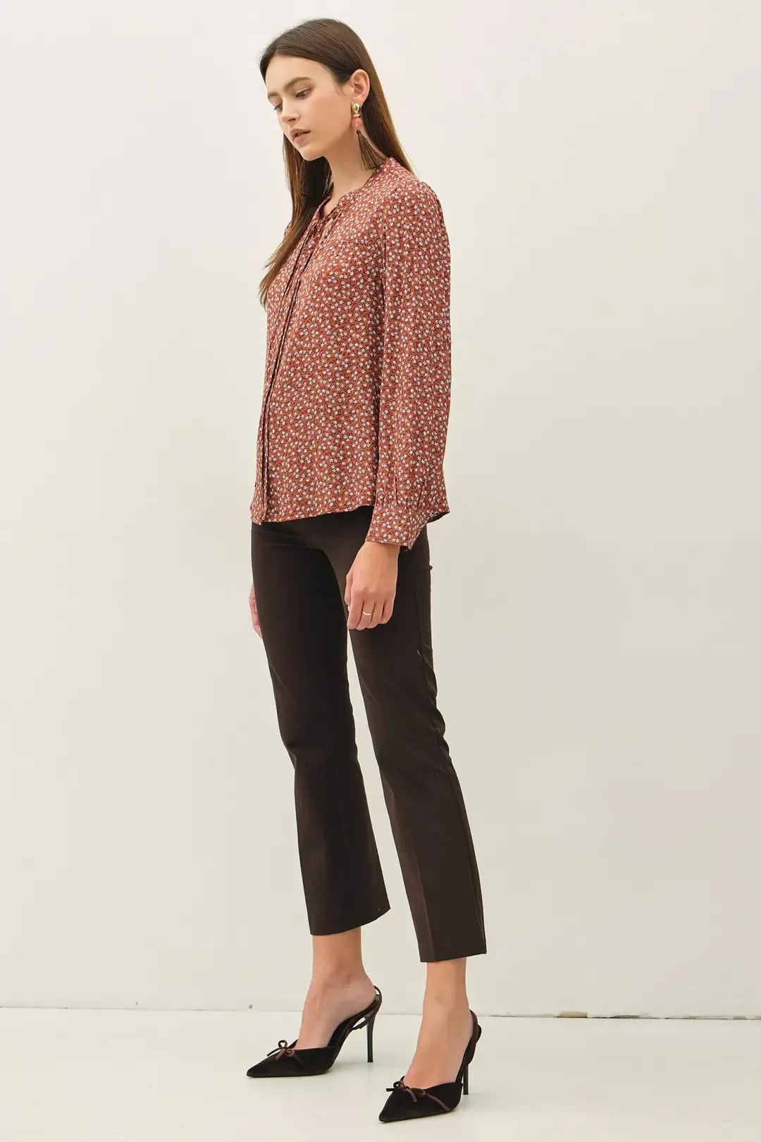 A woman wearing a rust-colored ditsy printed tie front collar blouse with long sleeves paired with black cropped trousers