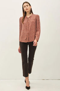 A woman wears a rust-colored ditsy printed tie front collar blouse with long sleeves paired with black cropped trousers