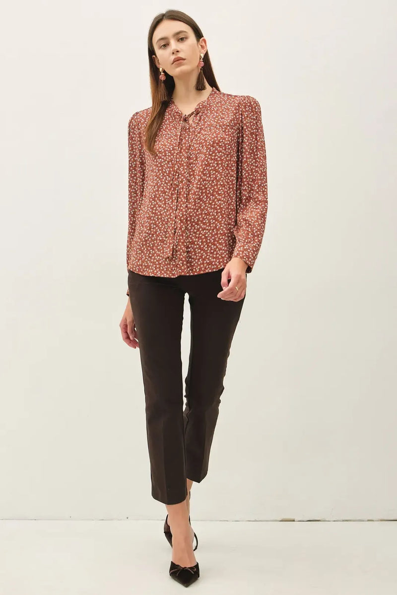 A woman wears a rust-colored ditsy printed tie front collar blouse with long sleeves paired with black cropped trousers