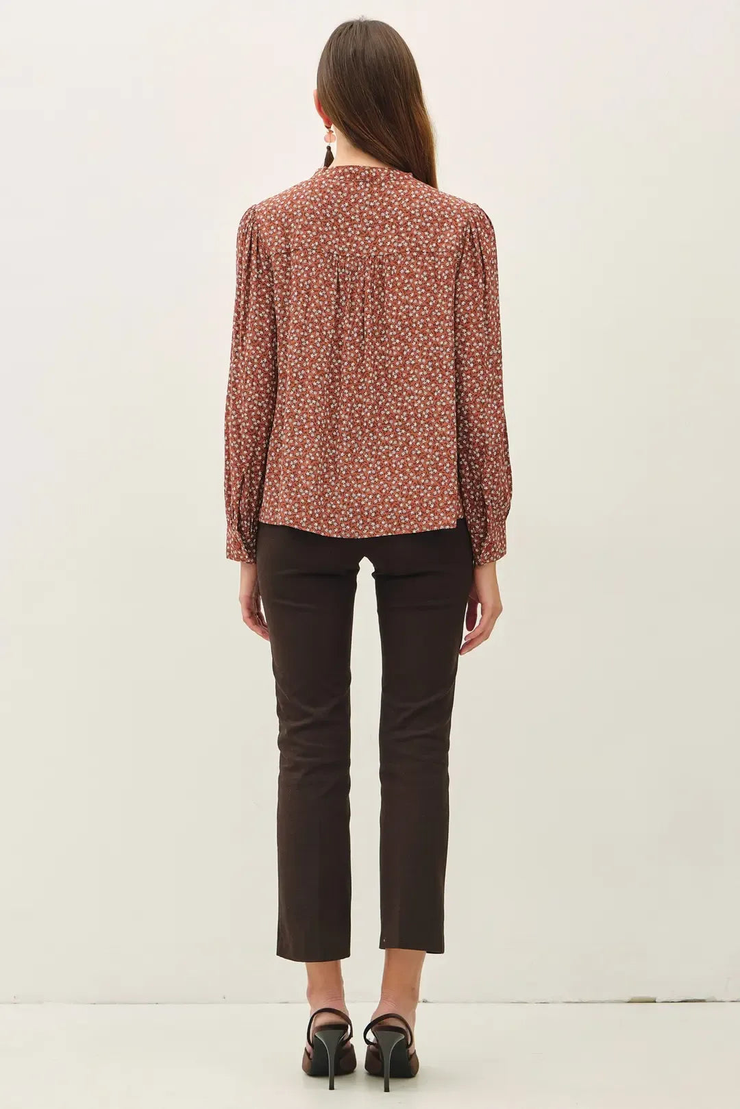 A woman wears a rust-colored ditsy printed tie front collar blouse with long sleeves paired with dark brown trousers
