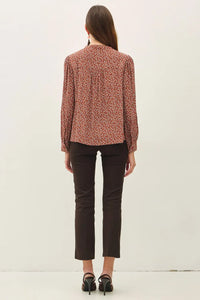 A woman wears a rust-colored ditsy printed tie front collar blouse with long sleeves paired with dark brown trousers