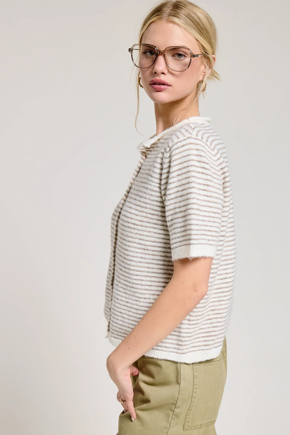 A woman wearing a striped short sleeve button cardigan in gray and white with cropped design and sleeve button detail