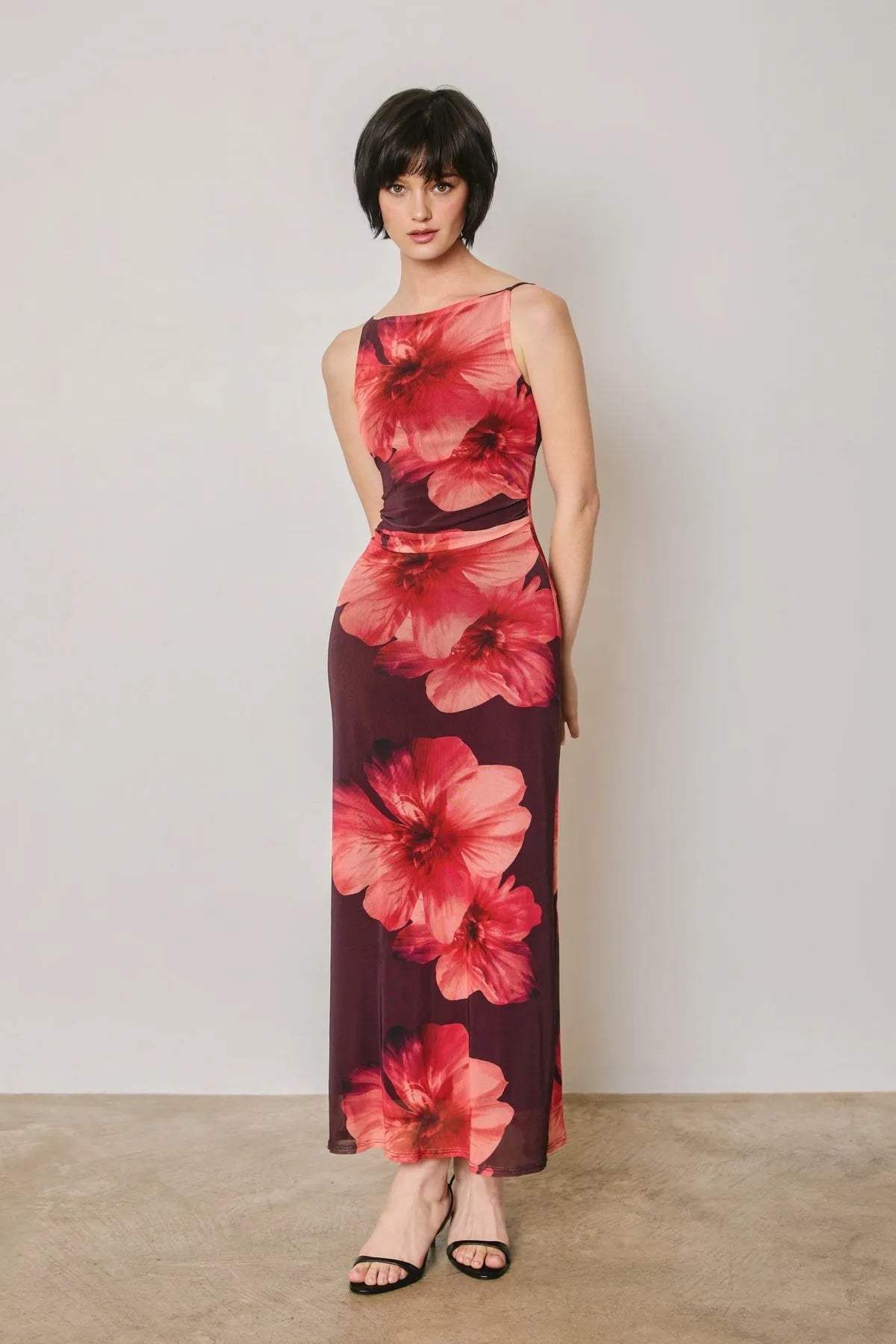 A woman wears a sleeveless floral mesh open back maxi dress with bold red floral patterns on deep burgundy fabric