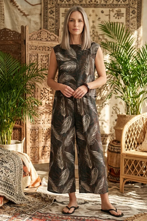 A woman wearing a sleeveless leaf print wide shoulder jumpsuit with a dark base and white leaf patterns