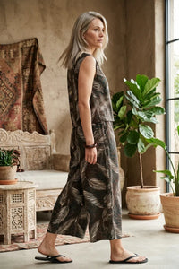 A woman wears a dark brown leaf print wide shoulder sleeveless jumpsuit with light beige leaf patterns
