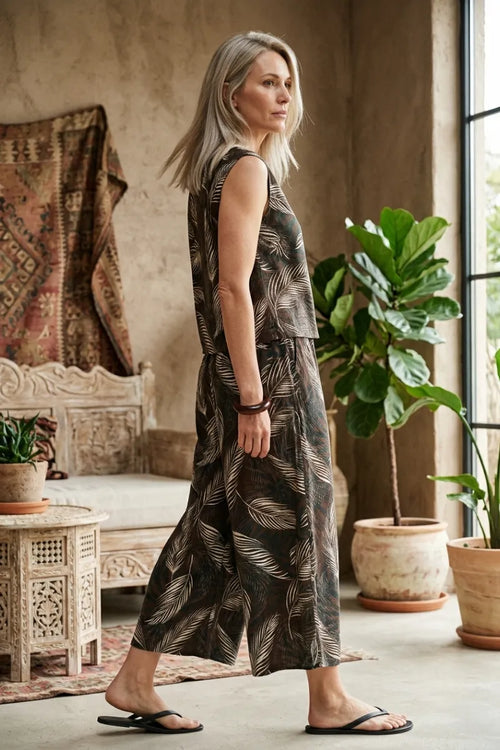 A woman wears a dark brown leaf print wide shoulder sleeveless jumpsuit with light beige leaf patterns